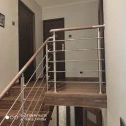 Nowoczesna balustrada ze stali nierdzewnej z drewnianym pochwytem, zamontowana na schodach wykończonych panelami imitującymi drewno, widok na dwie ciemne, wewnętrzne drzwi.