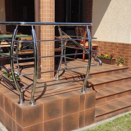 Spawstal-balustrady nierdzewne - Zewnętrzne schody wejściowe wyłożone brązowymi płytkami, zabezpieczone metalową, chromowaną konstrukcją z poziomych prętów. Dodatkowa poręcz przy ścianie. Na schodach doniczki z kwiatami.