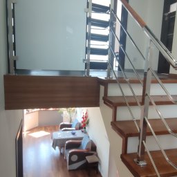 Spawstal-balustrady nierdzewne - Wnętrze domu z drewnianymi schodami z metalową balustradą i poręczą, z widoczną szklaną balustradą na antresoli i salonem z drewnianą podłogą w tle.