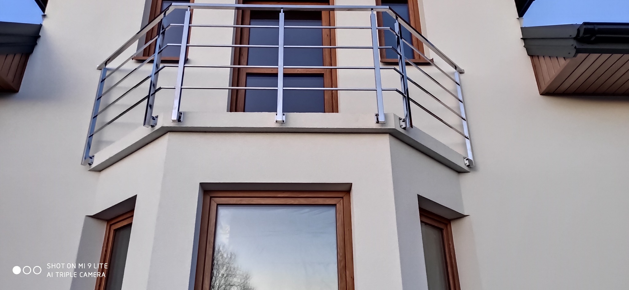 Nowoczesna, metalowa balustrada balkonowa z polerowanej stali, zamontowana na jasnej elewacji budynku z drewnianą stolarką okienną.