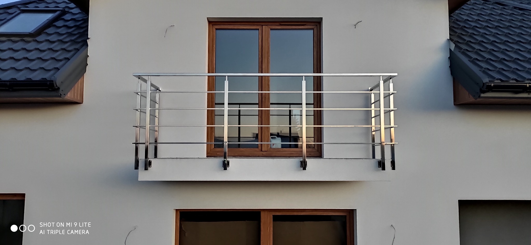 Balkon z prostą, chromowaną barierką z poziomych rurek, widok z dołu na tle białej elewacji i drewnianej ramy okiennej.