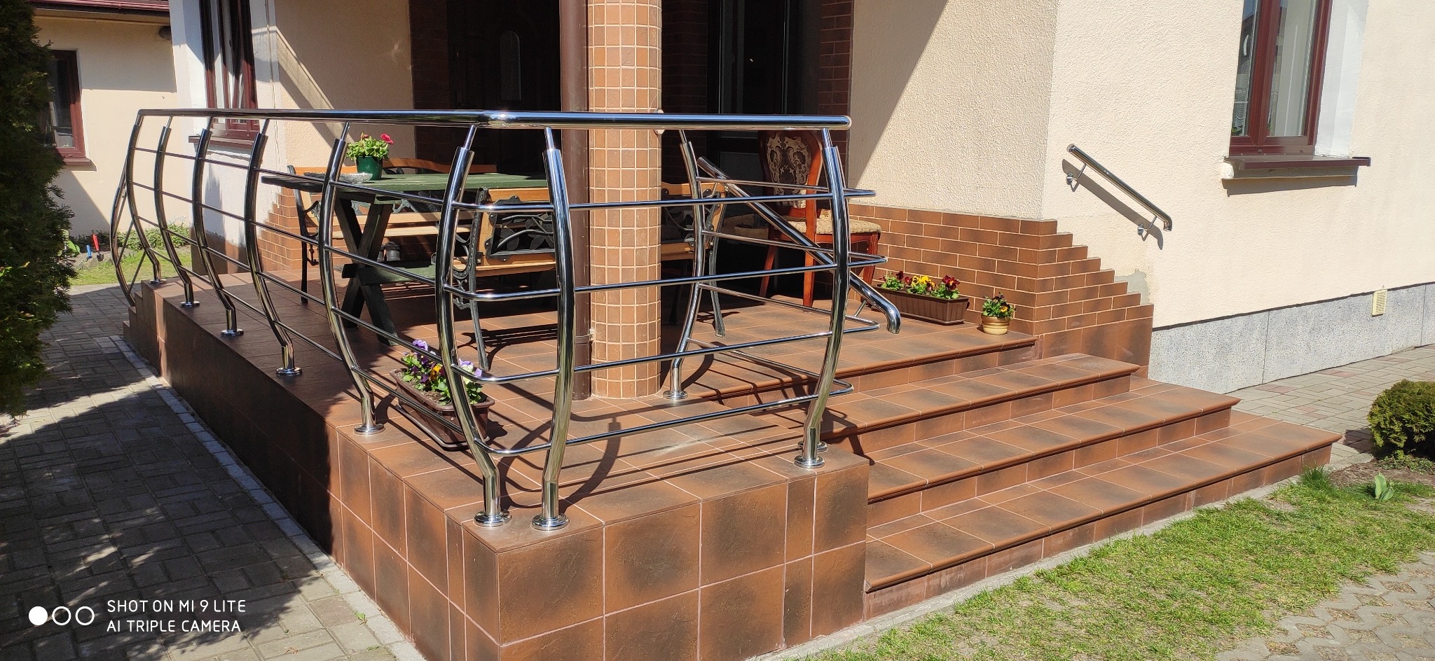 Zewnętrzne schody wejściowe wyłożone brązowymi płytkami, zabezpieczone metalową, chromowaną konstrukcją z poziomych prętów. Dodatkowa poręcz przy ścianie. Na schodach doniczki z kwiatami.