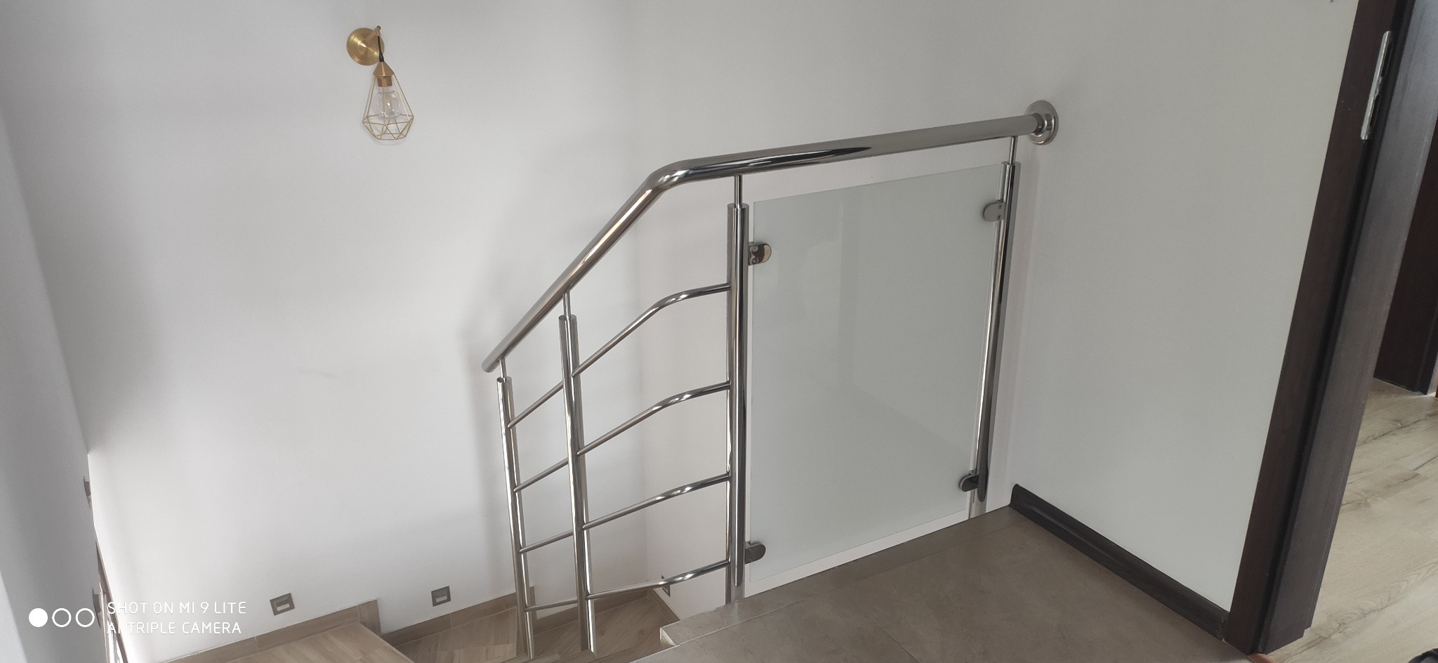 Schody z balustradą ze stali nierdzewnej i szkła, widok z góry na stopnie wyłożone płytkami, na ścianie lampa w stylu industrialnym.