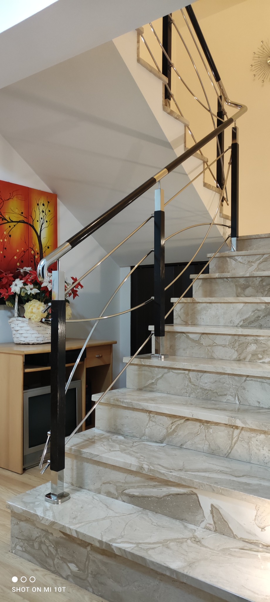 Marmurowe schody z nowoczesną balustradą ze stali nierdzewnej i elementami z ciemnego drewna, w tle obraz i kwiaty.