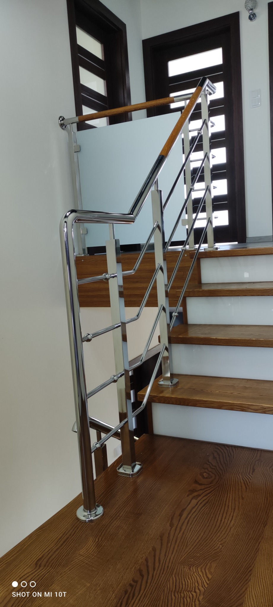 Wewnętrzne schody z drewnianymi stopniami i chromowanymi balustradami z wypełnieniem z mlecznego szkła, widoczne drzwi w tle.