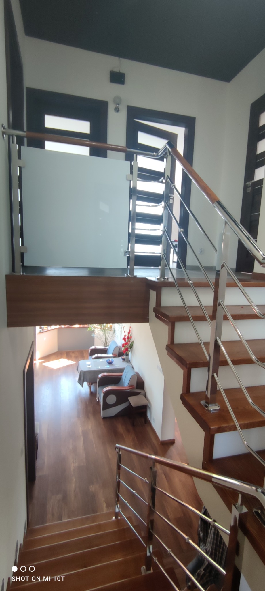 Wnętrze domu z drewnianymi schodami z metalową balustradą i poręczą, z widoczną szklaną balustradą na antresoli i salonem z drewnianą podłogą w tle.