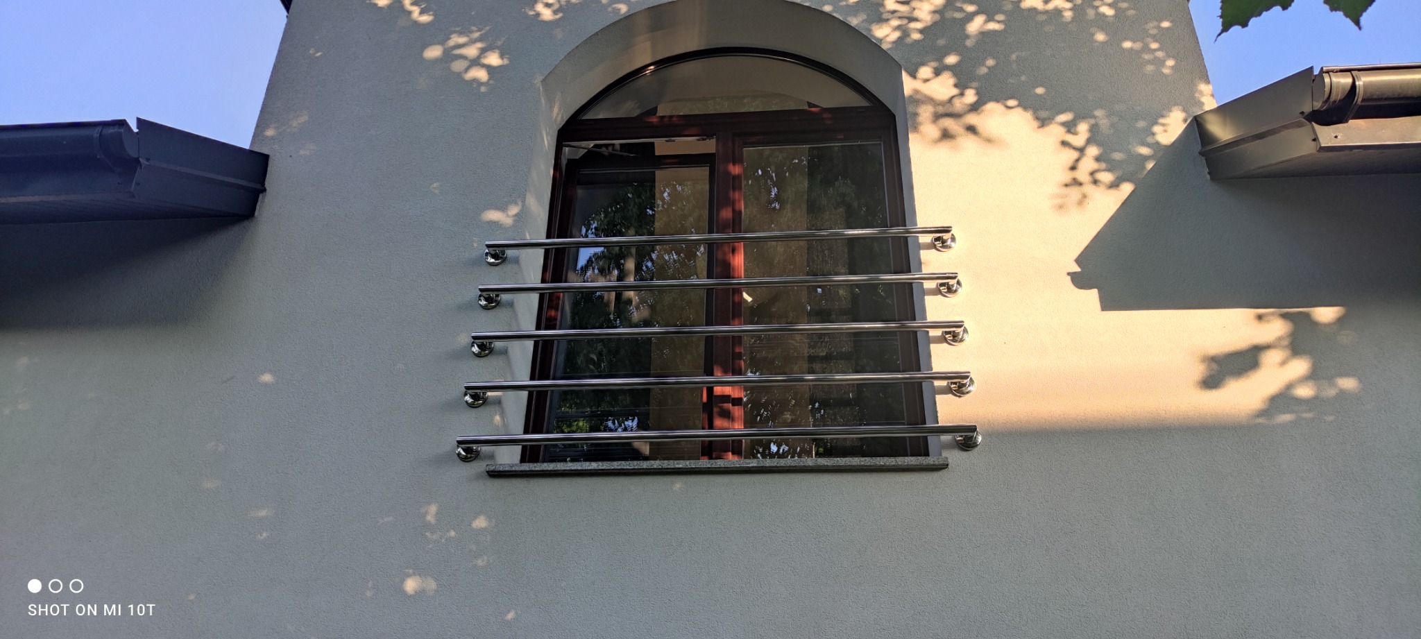 Nierdzewna balustrada okienna z czterema poziomymi prętami i zaokrąglonymi zakończeniami, zamontowana na elewacji budynku z oknem łukowym.