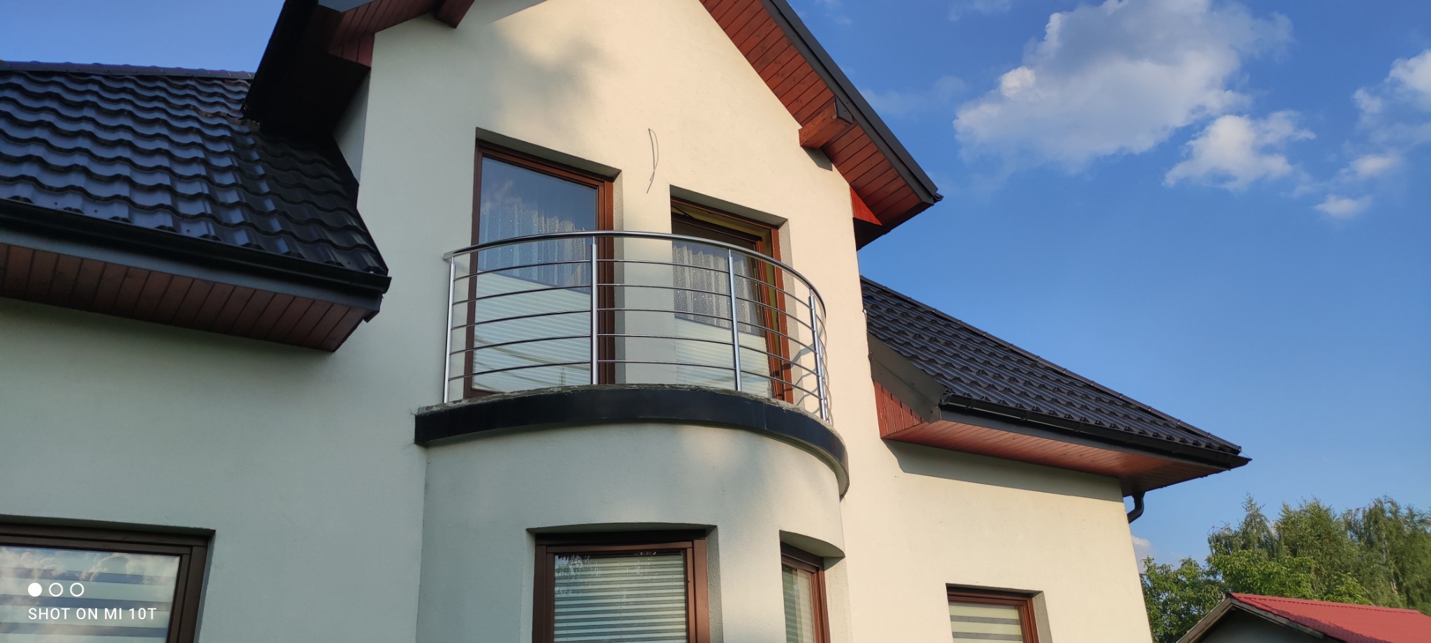 Biały dom z balkonem zabezpieczonym nowoczesną, metalową balustradą z poziomych rurek, na tle błękitnego nieba z chmurami.