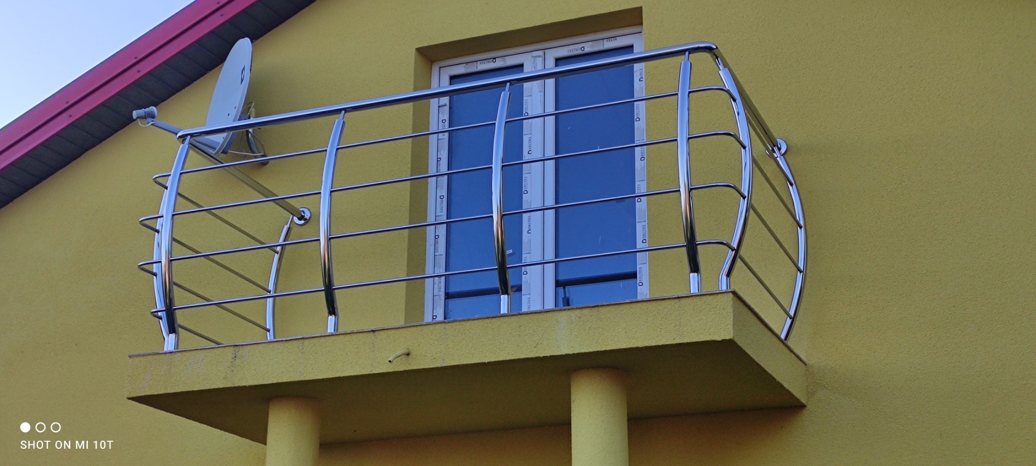 Balkon z chromowaną, giętą balustradą z rurek, na tle żółtej elewacji budynku z białą stolarką okienną i anteną satelitarną.