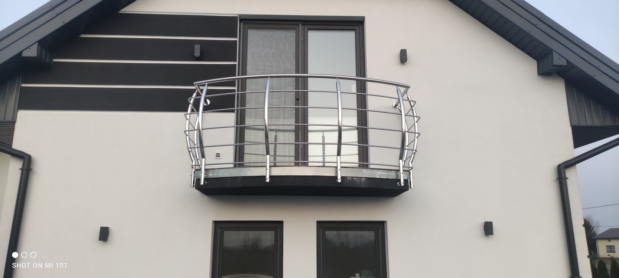 Nowoczesny balkon z balustradą ze stali nierdzewnej i szklanym wypełnieniem, zamontowany na elewacji domu z białym tynkiem i czarnymi akcentami.