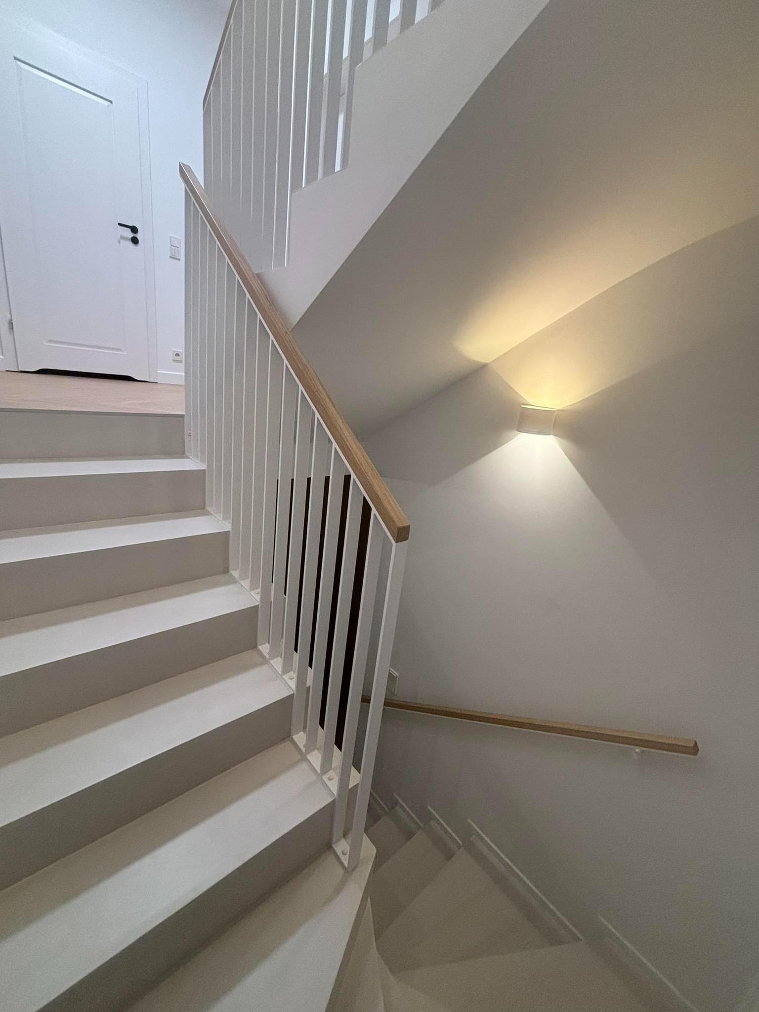 Nowoczesne, białe schody z drewnianą poręczą i minimalistyczną, białą balustradą z pionowych słupków, oświetlone kinkietem ściennym, widok z dołu.