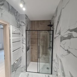 Dom-tech - Wykończona łazienka z kabiną prysznicową z czarnymi ramami, grzejnikiem drabinkowym i płytkami imitującymi marmur na ścianach i podłodze, oraz drewnopodobnymi płytkami wewnątrz kabiny.