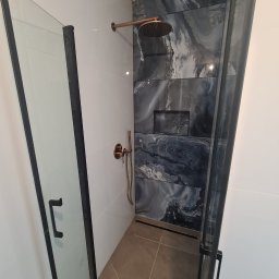 Dom-tech - Nowoczesna kabina prysznicowa z czarnymi profilami i drzwiami ze szkła hartowanego, ściana wyłożona płytkami imitującymi marmur w odcieniach szarości i bieli, armatura w kolorze miedzi.