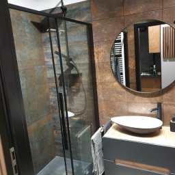 Dom-tech - Nowoczesna łazienka z kabiną prysznicową typu walk-in z czarną ramą, umywalką nablatową na szafce z drewnianym blatem i lustrem w okrągłej ramie na tle ściany wyłożonej płytkami imitującymi rdzę.