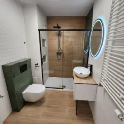 Nowoczesna aranżacja łazienki w Poznaniu z drewnopodobnymi płytkami podłogowymi i ściennymi w kabinie prysznicowej, białą armaturą, podświetlanym lustrem i zielonym elementem zabudowy toalety.