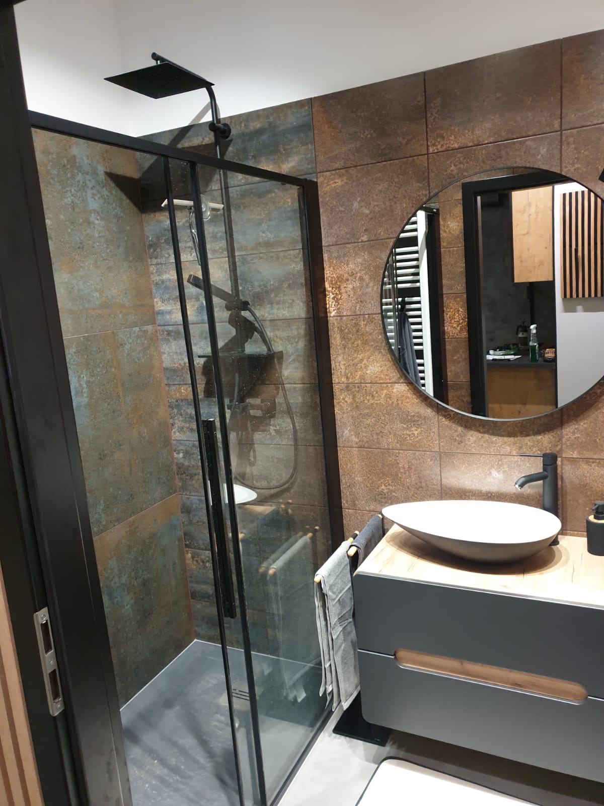Nowoczesna łazienka z kabiną prysznicową typu walk-in z czarną ramą, umywalką nablatową na szafce z drewnianym blatem i lustrem w okrągłej ramie na tle ściany wyłożonej płytkami imitującymi rdzę.