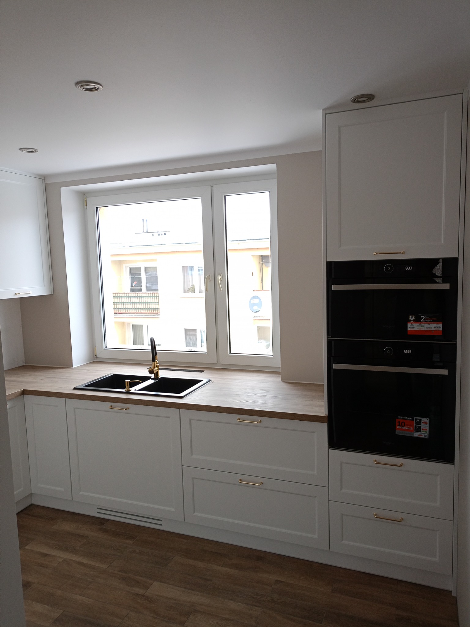 Nowoczesna, jasna kuchnia z białymi szafkami, drewnianym blatem i czarnym zlewem z złotą armaturą, oświetlona światłem dziennym wpadającym przez okno.