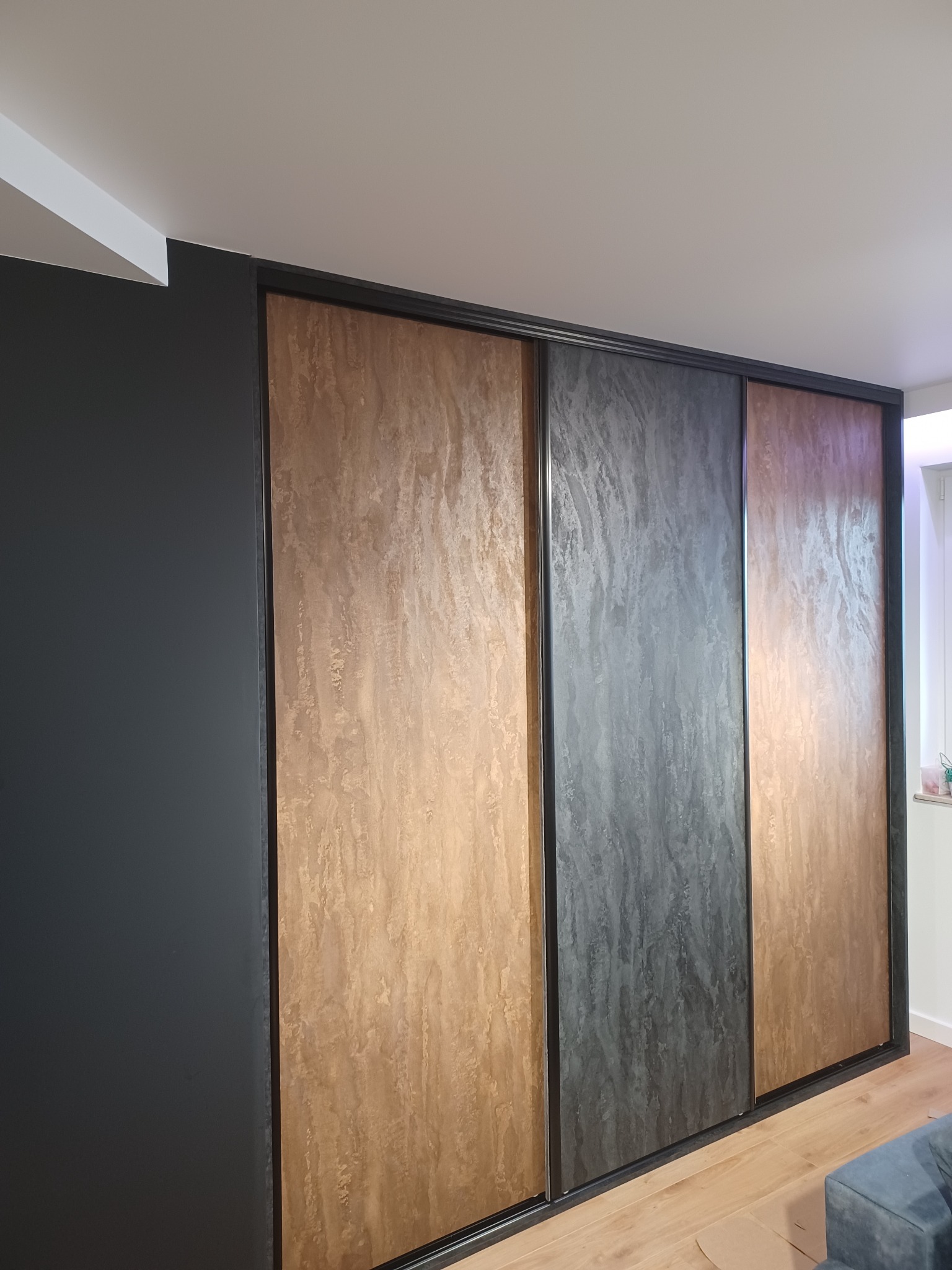 Nowoczesna szafa wnękowa z przesuwnymi drzwiami o teksturze drewna i kamienia, wkomponowana w ciemną ścianę, widok z dołu.