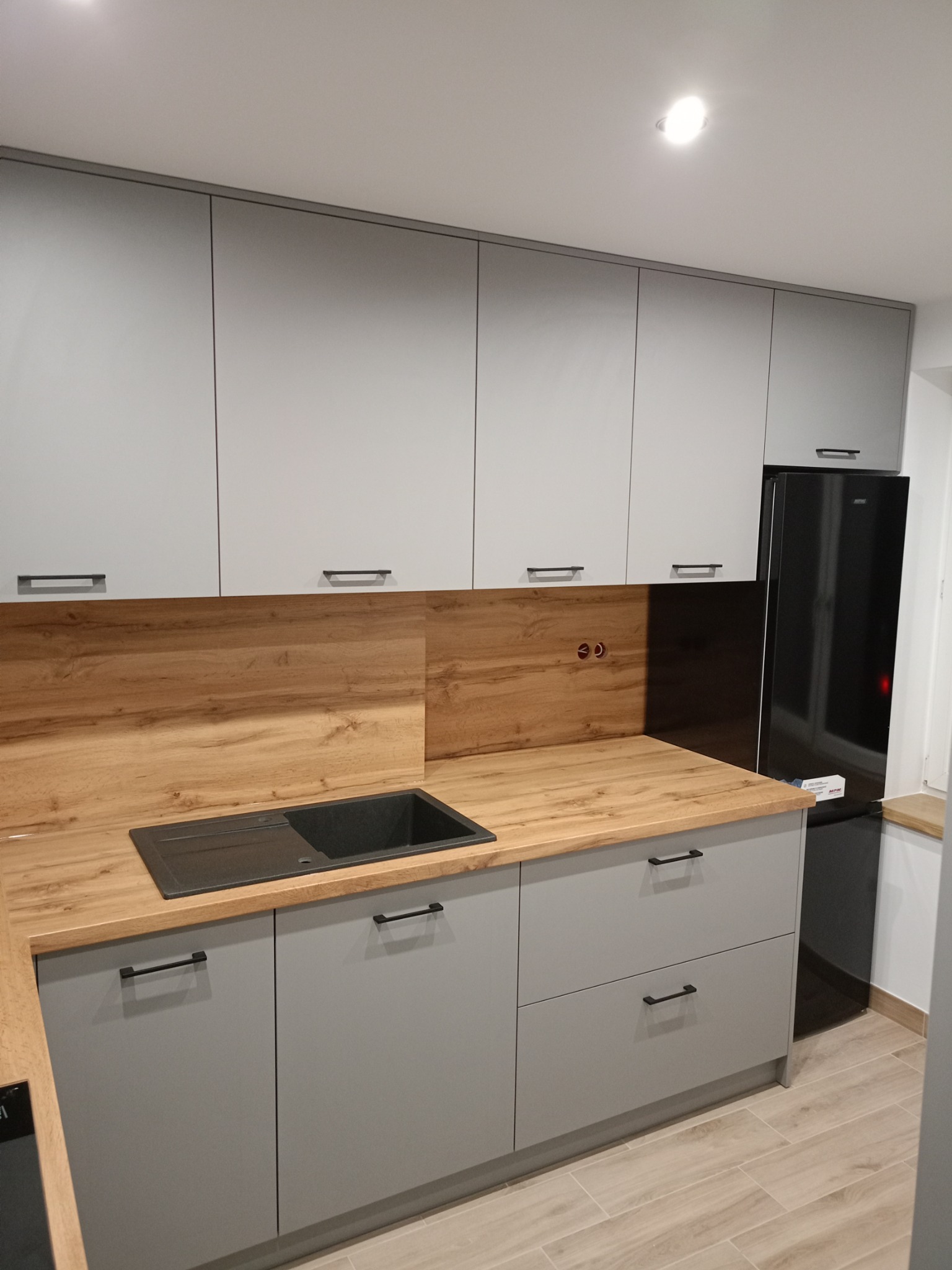 Szara zabudowa kuchenna z drewnopodobnym blatem i czarnym zlewem, widoczne gniazdka elektryczne na ścianie, minimalistyczne uchwyty i czarna lodówka w tle.