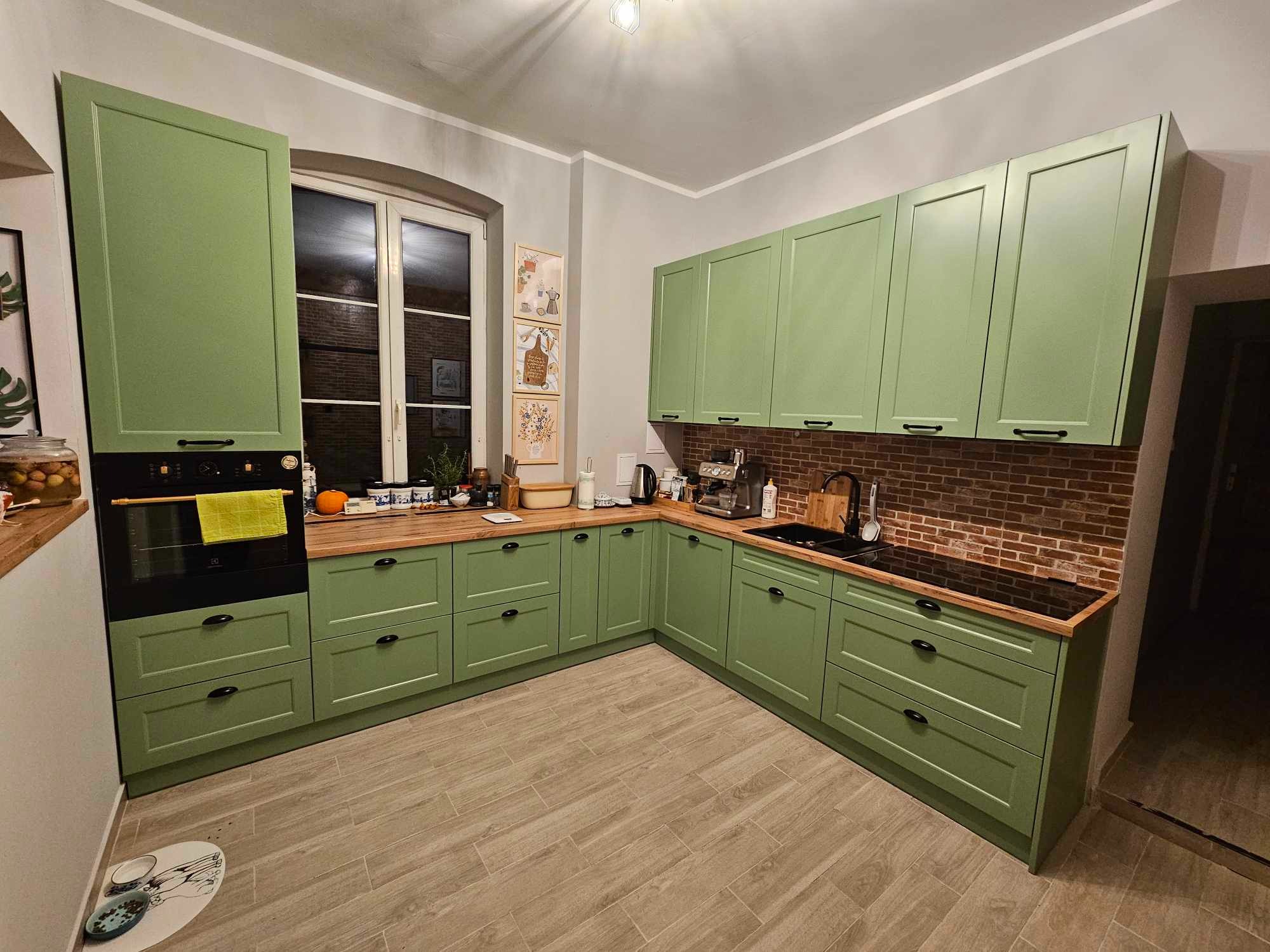 Jasnozielona, narożna zabudowa kuchenna z drewnianym blatem, czarnym piekarnikiem i płytą grzewczą, oraz ceglaną ścianą nad blatem, oświetlona ciepłym światłem.