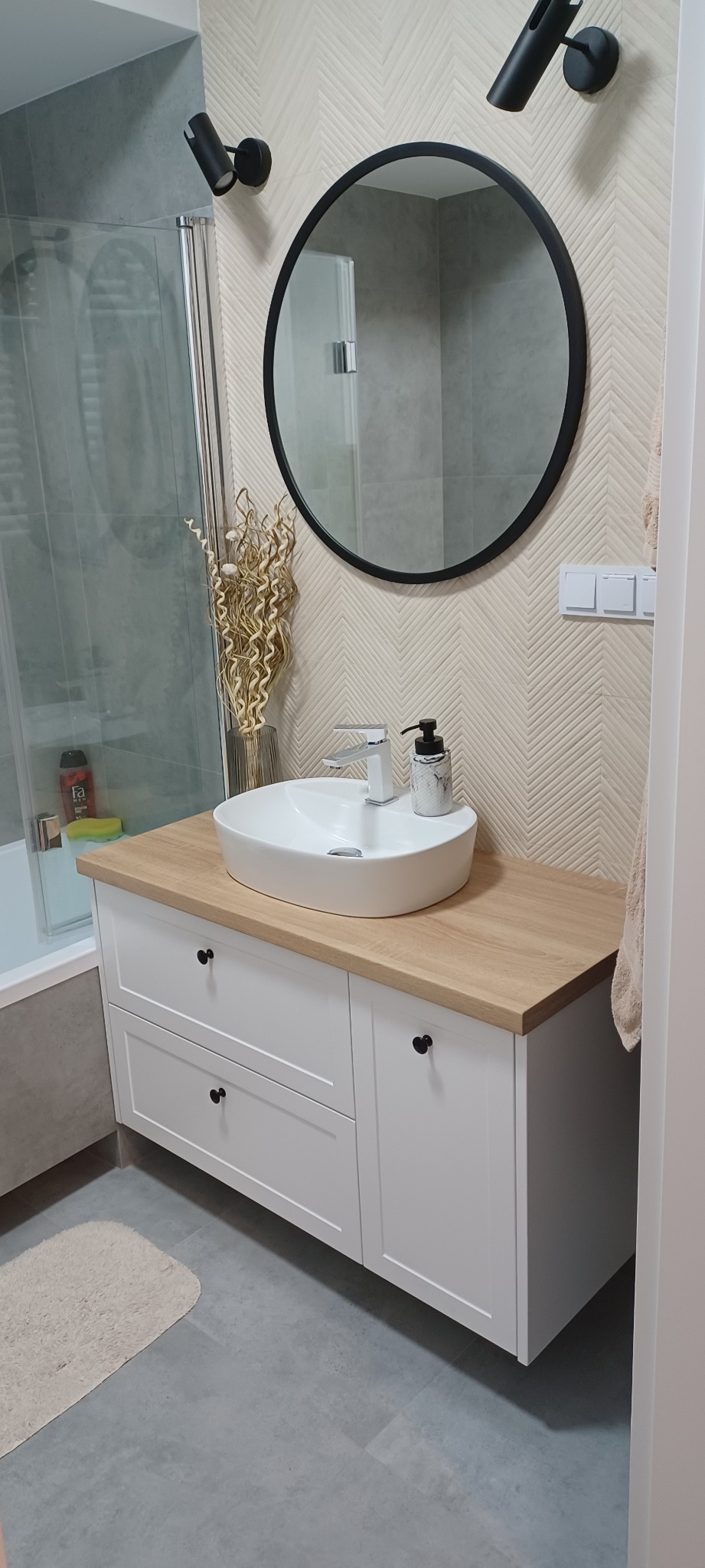 Biała szafka łazienkowa z drewnianym blatem, owalna umywalka nablatowa, okrągłe lustro w czarnej ramie i dekoracyjne oświetlenie punktowe w łazience z beżową tapetą w jodełkę.