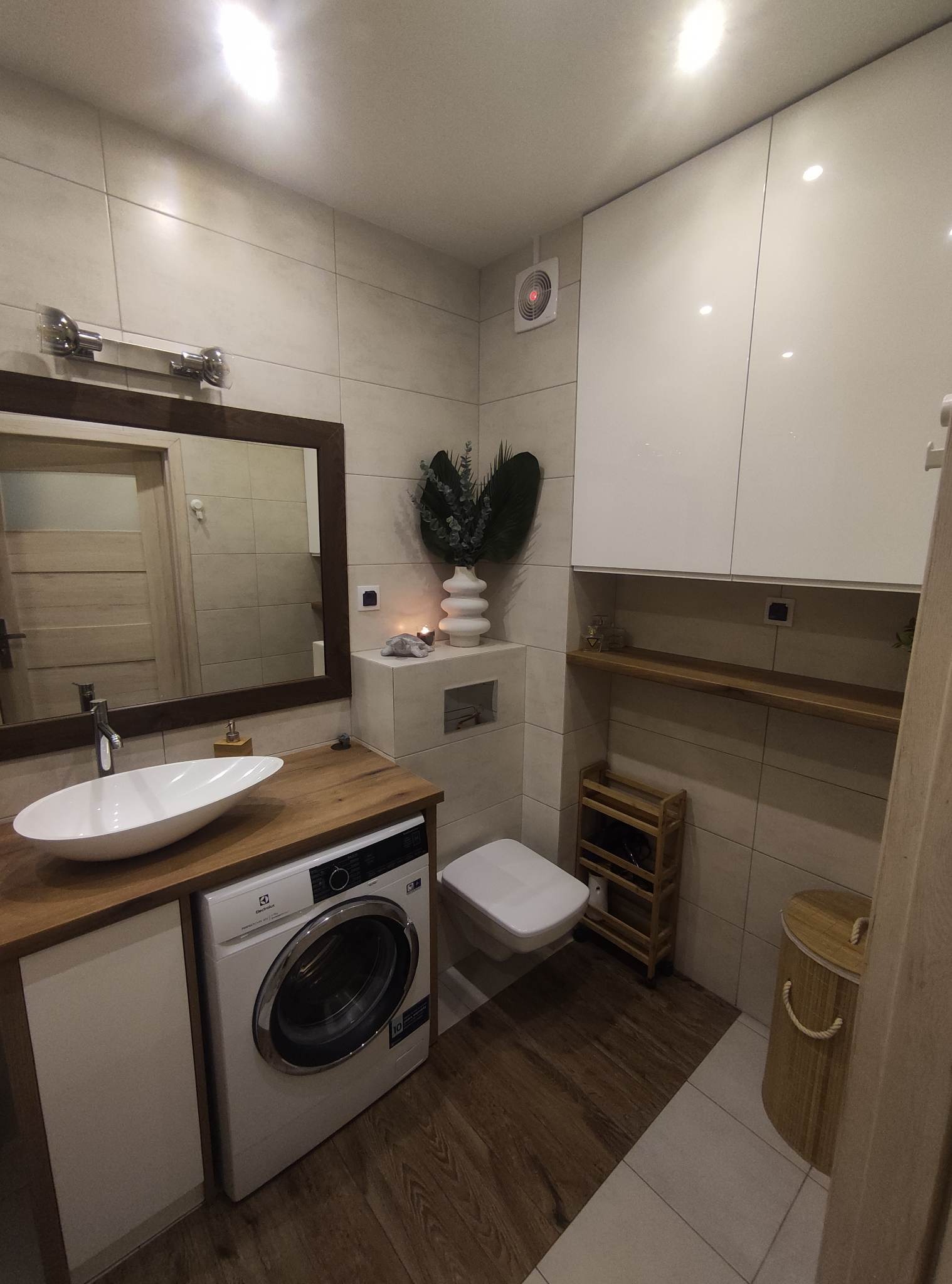 Nowoczesna łazienka z drewnianym blatem, umywalką nablatową, pralką Electrolux i szafkami wiszącymi z białymi frontami, uzupełniona o drewniane akcenty i dekoracje.