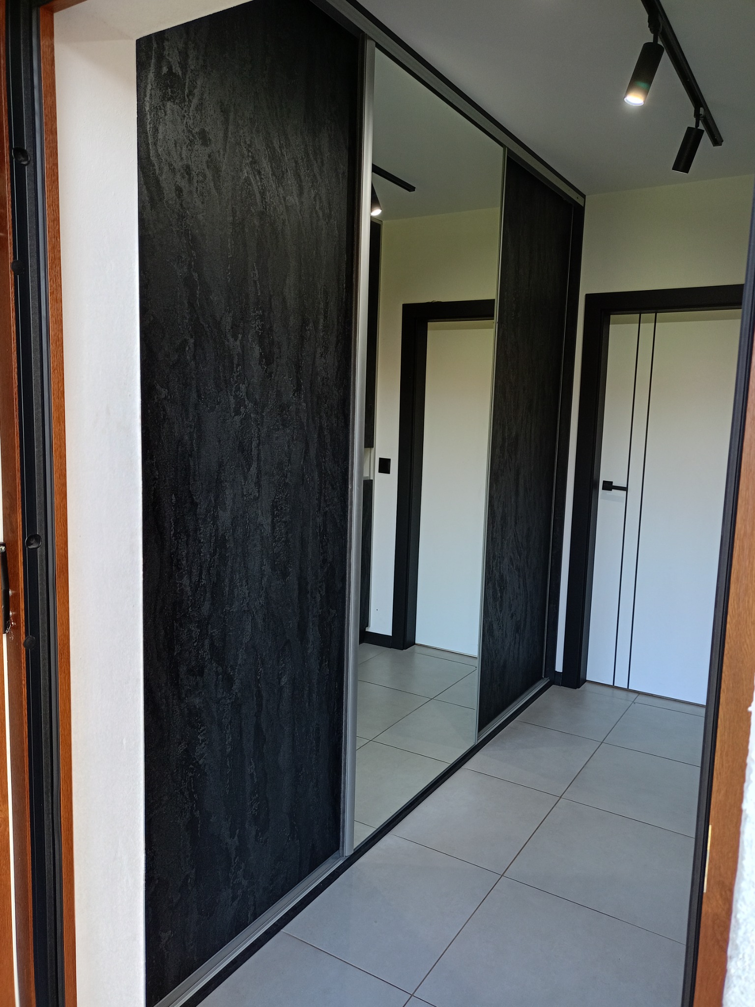 Wnętrze przedpokoju z zabudowaną szafą przesuwną z czarnymi, teksturowanymi drzwiami i lustrem, odbijającym białe drzwi z czarnymi ramami.