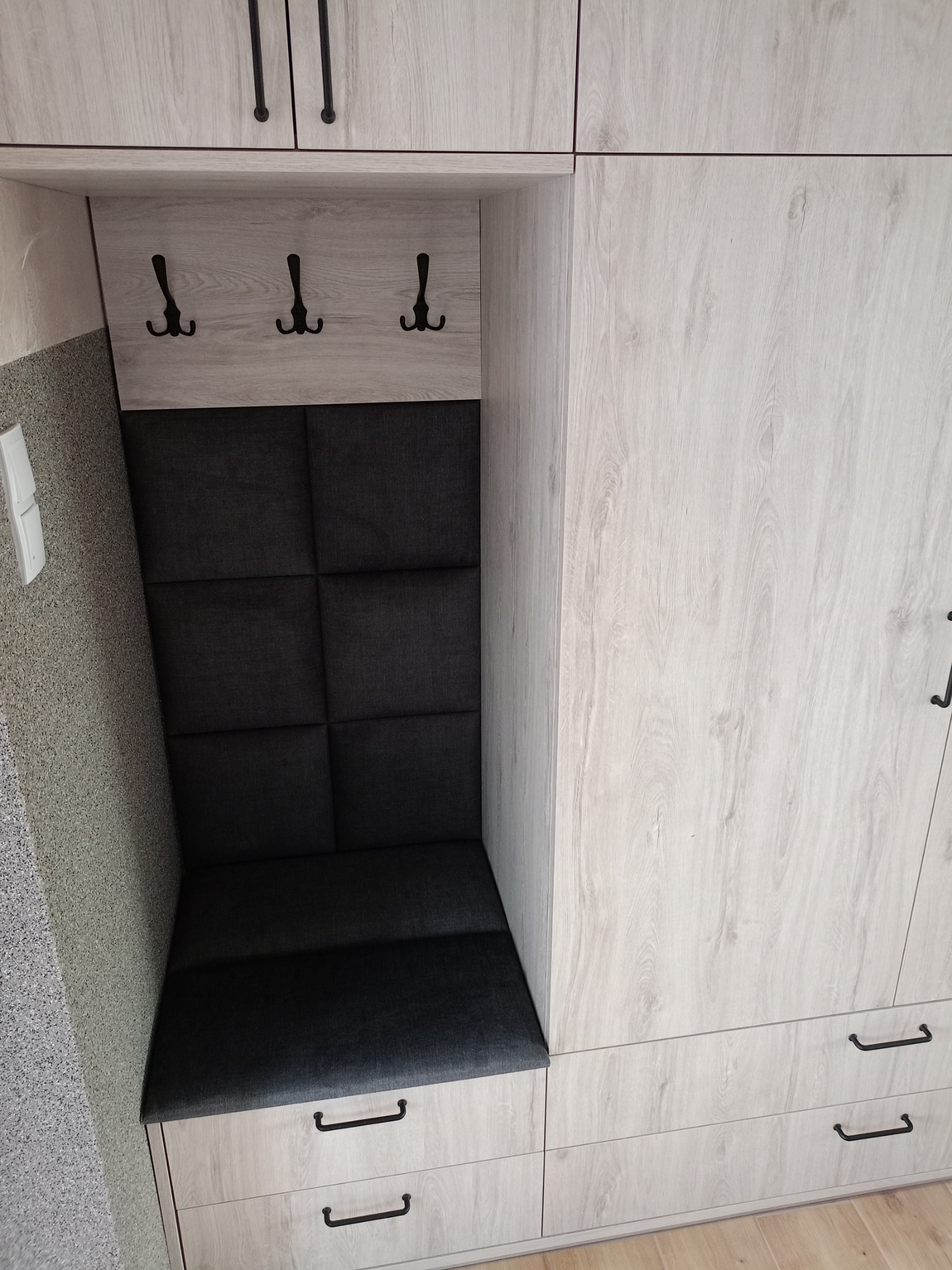 Zabudowa przedpokoju z siedziskiem i wieszakami, fronty w jasnym odcieniu drewna, czarne uchwyty i tapicerowane panele na ścianie.