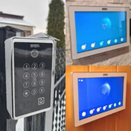 Zestaw wideodomofonowy STECH: panel zewnętrzny z klawiaturą numeryczną zamontowany na metalowej bramie w deszczowy dzień oraz panel wewnętrzny z ekranem dotykowym pokazującym datę i godzinę...