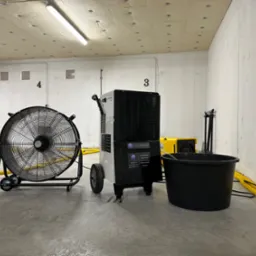 Profesjonalny osuszacz powietrza Trotec, wentylator przemysłowy i czarny pojemnik na wodę w surowym, betonowym pomieszczeniu z ż&oacute;łtymi wężami, prawdopodobnie garażu podziemnym.