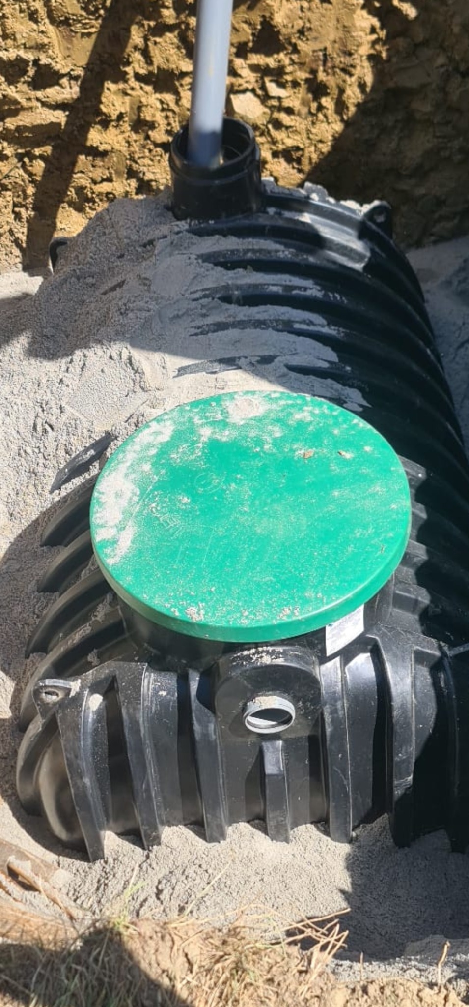 Częściowo zasypany piaskiem czarny, karbowany zbiornik z zieloną pokrywą i wystającą rurą kanalizacyjną, umieszczony w wykopie ziemnym.