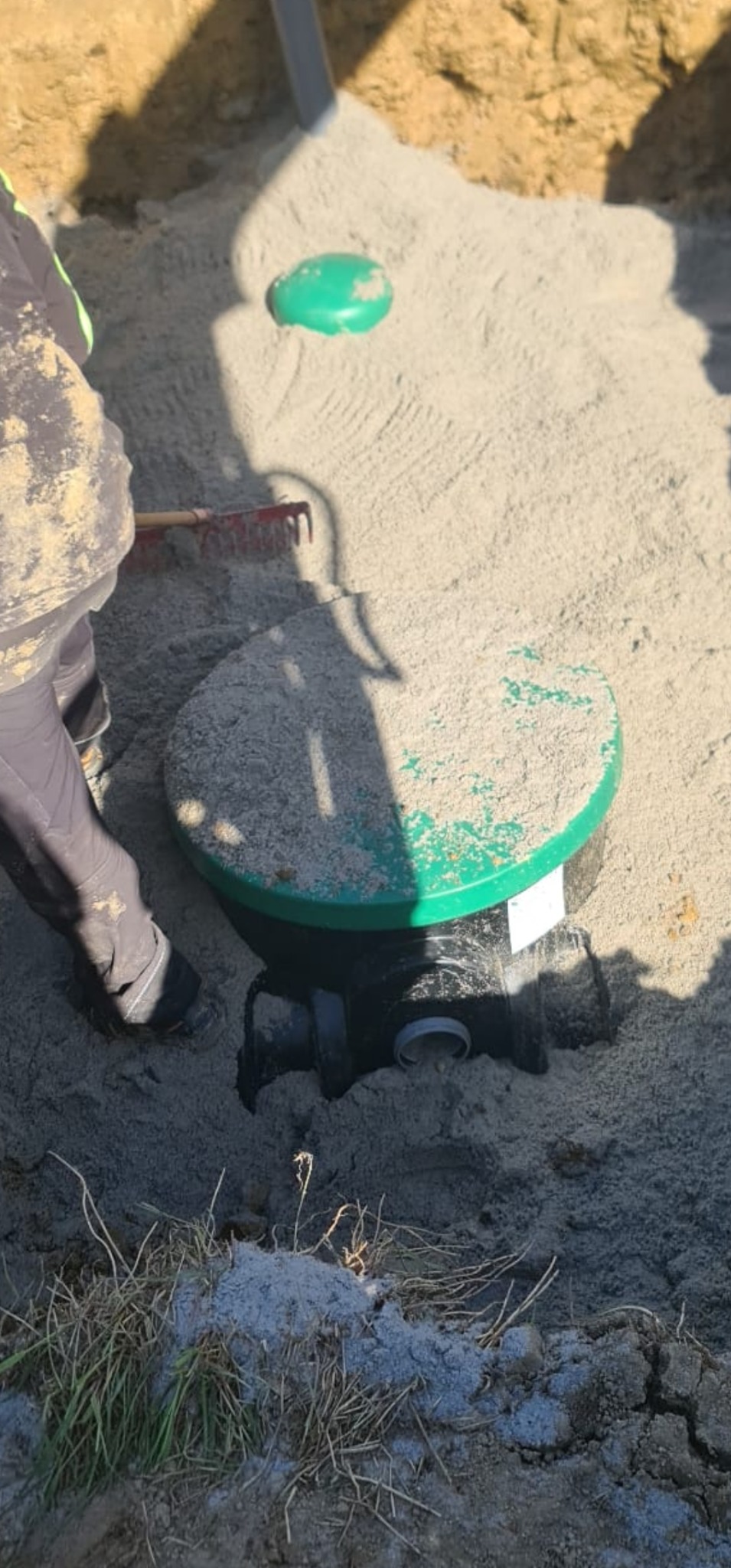 Montaż zielonego zbiornika na szambo, częściowo zasypanego piaskiem, z widocznymi rurami podłączeniowymi i pracownikiem wyrównującym teren grabiami.