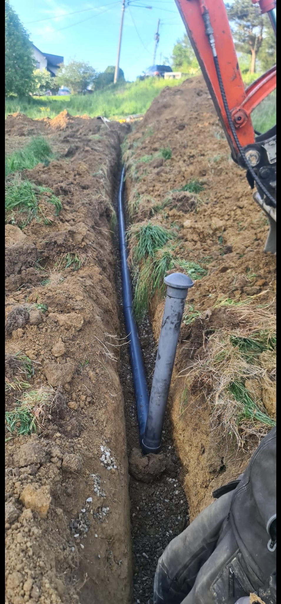 Wykopany wąski rów z ułożonymi rurami drenażowymi, częściowo zasypany żwirem, widoczny fragment koparki, trawa na skraju wykopu.