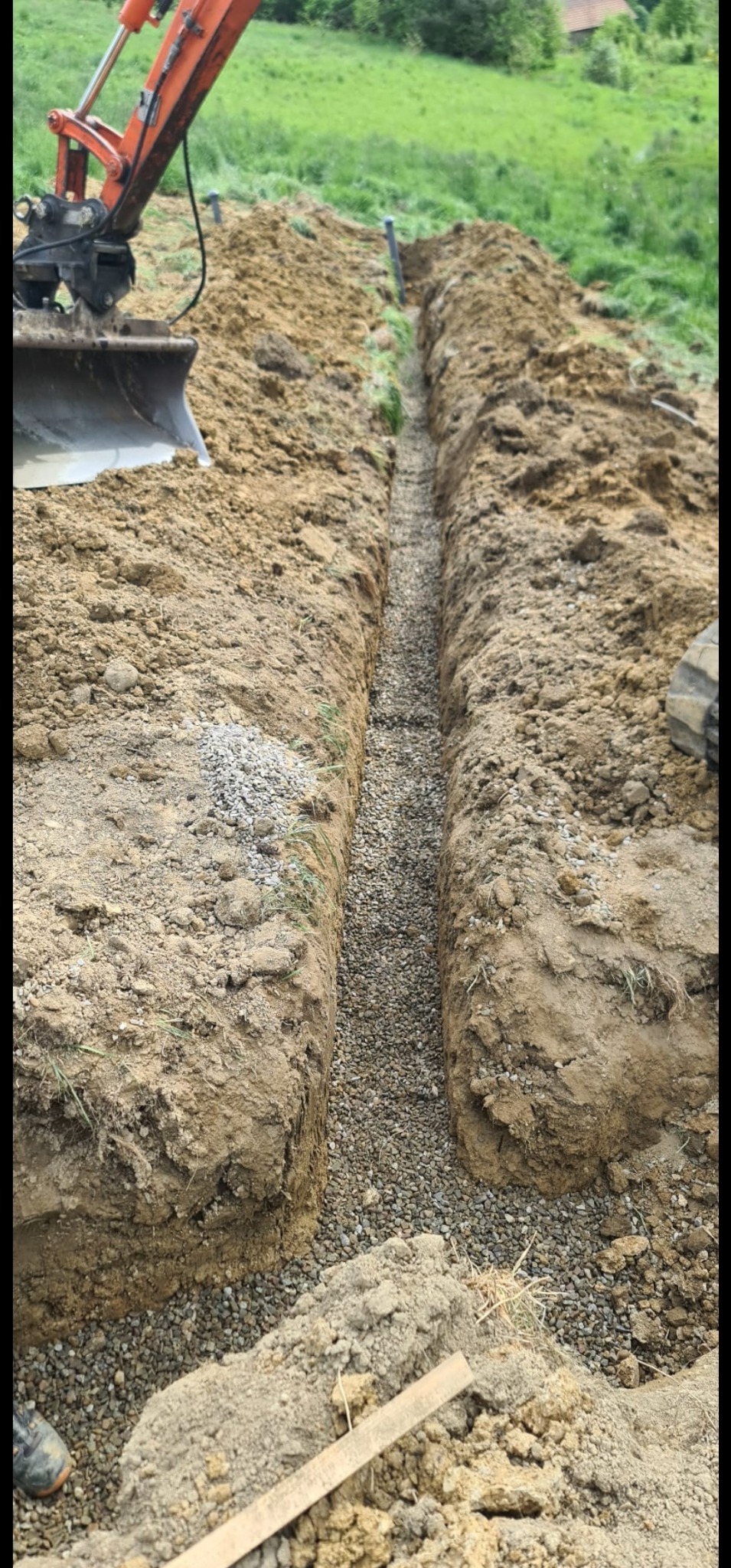 Wykopany wąski rów wypełniony szarym żwirem, z widoczną pomarańczową koparką w tle i fragmentem buta roboczego w dolnym rogu.