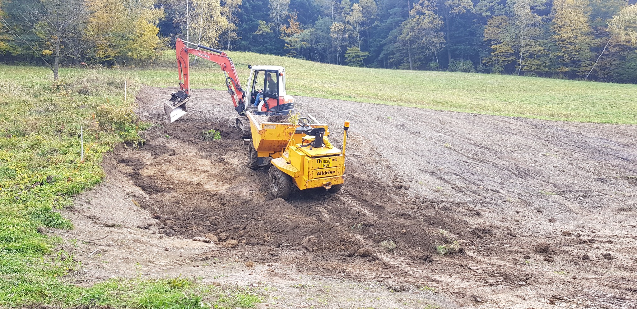 Czerwono-biała minikoparka Kubota i żółta wywrotka AllDrive na nierównym, błotnistym terenie z kępami trawy i drzewami w tle, wskazujące na prace ziemne.
