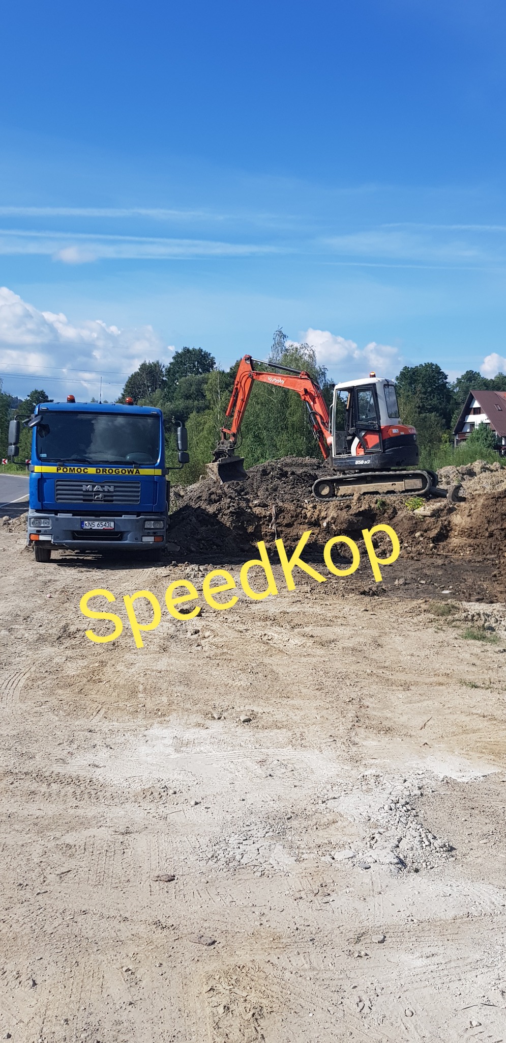 Pomarańczowo-biała koparka Kubota wykonuje prace ziemne na placu budowy, obok stoi niebieska ciężarówka pomocy drogowej MAN z włączonymi światłami ostrzegawczymi, w tle drzewa i niebo.