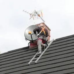 Pracownik na drabinie, zabezpieczony uprzężą, instaluje antenę satelitarną na kominie, z widoczną anteną naziemną powyżej, na tle pochmurnego nieba.