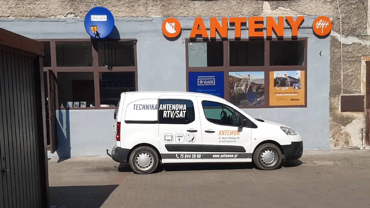 Biały samochód dostawczy z napisem 'Technika Antenowa RTV/SAT' zaparkowany przed budynkiem firmy z szyldem 'Anteny' w kolorze pomarańczowym i logiem anteny.