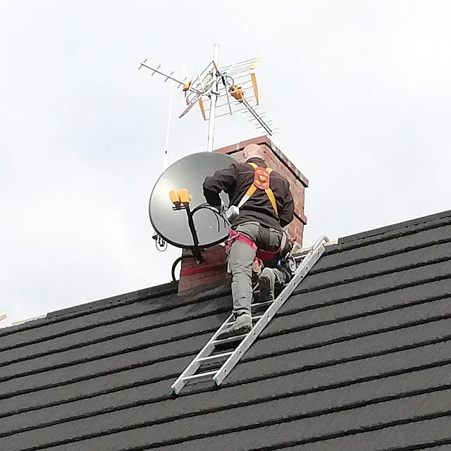 Pracownik na drabinie, zabezpieczony uprzężą, instaluje antenę satelitarną na kominie, z widoczną anteną naziemną powyżej, na tle pochmurnego nieba.