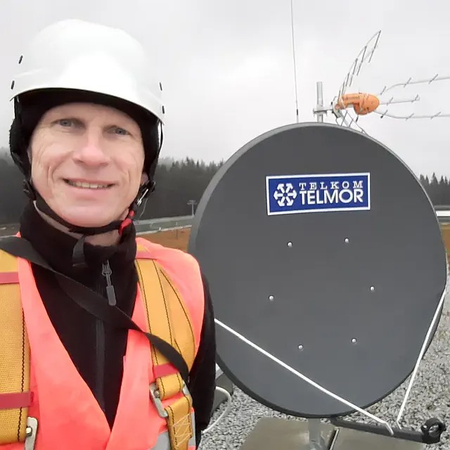 Uśmiechnięty monter w kasku i kamizelce odblaskowej pozuje obok szarej anteny satelitarnej Telmor i anteny naziemnej na tle pochmurnego nieba i drzew.