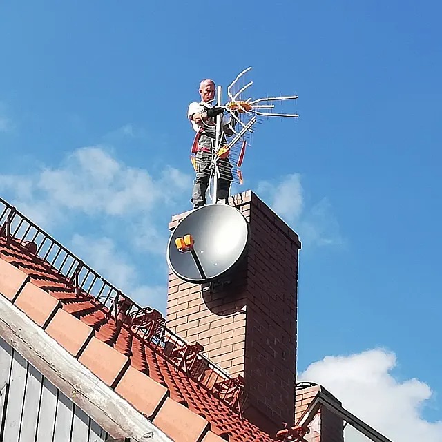 Pracownik w uprzęży na kominie, instaluje antenę Yagi, widoczna antena satelitarna, dachówka ceramiczna i błękitne niebo.