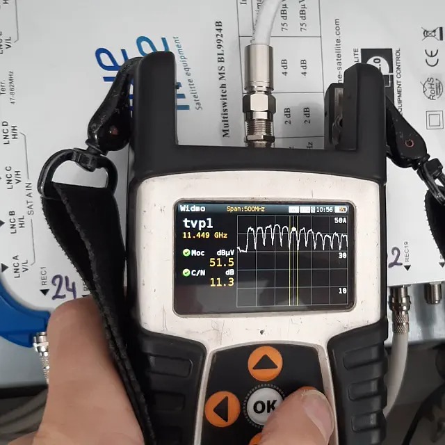 Pomiar sygnału antenowego za pomocą miernika w trakcie montażu, widoczny ekran z wykresem częstotliwości i wartościami mocy w dBμV, kabel antenowy podłączony do multiswitcha MS BL9924B.