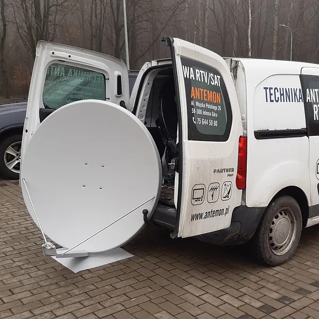 Biała antena satelitarna wystająca z otwartych drzwi bagażnika białego samochodu dostawczego z logo firmy i danymi kontaktowymi na karoserii, zaparkowanego na brukowanym placu.