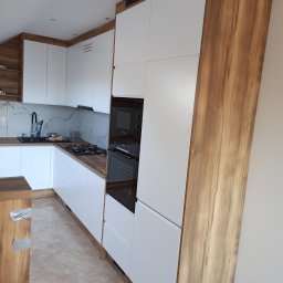 Krzysztof Gawlak - Nowoczesna kuchnia z białymi frontami i drewnianymi akcentami, zabudowa kuchenna z piekarnikiem i płytą gazową, marmurowy backsplash.