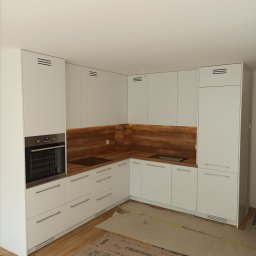 Krzysztof Gawlak - Nowoczesna, biała kuchnia z drewnianym blatem i panelem ściennym, z piekarnikiem w zabudowie i płytą indukcyjną, podłoga częściowo zabezpieczona kartonem.