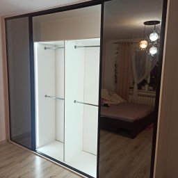 Krzysztof Gawlak - Wnętrze szafy przesuwnej z lustrzanymi drzwiami, widoczne półki i drążki na ubrania, odbicie pokoju z łóżkiem i lampą.