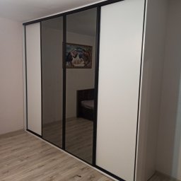 Krzysztof Gawlak - Szafa wnękowa z przesuwnymi drzwiami: białe panele boczne i lustrzane panele środkowe z czarnymi ramami, odbijające obraz na ścianie i część łóżka, na podłodze panele drewnopodobne.