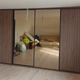 Trzydrzwiowa szafa przesuwna z lustrami i ciemnobrązowymi panelami drewnopodobnymi, odbijająca zagracony pokój z beżową sofą i workiem na podłodze.