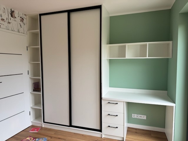 Biała szafa wnękowa z przesuwnymi drzwiami i czarnymi obramowaniami, połączona z biurkiem z szufladami i półkami wiszącymi na zielonej ścianie.