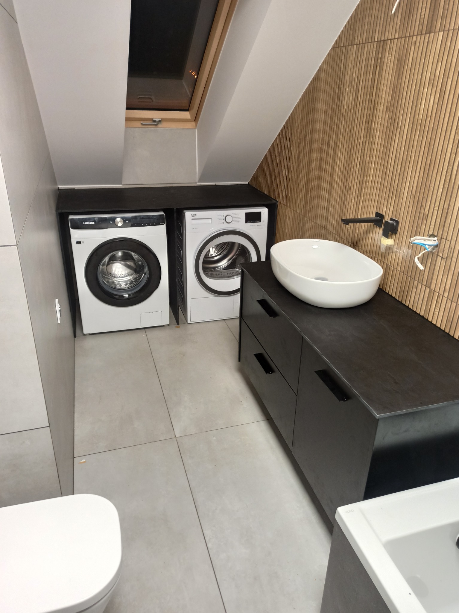 Łazienka na poddaszu z dwiema pralkami obok szafki umywalkowej z owalną umywalką nablatową, drewniana ściana z ozdobnymi listwami.